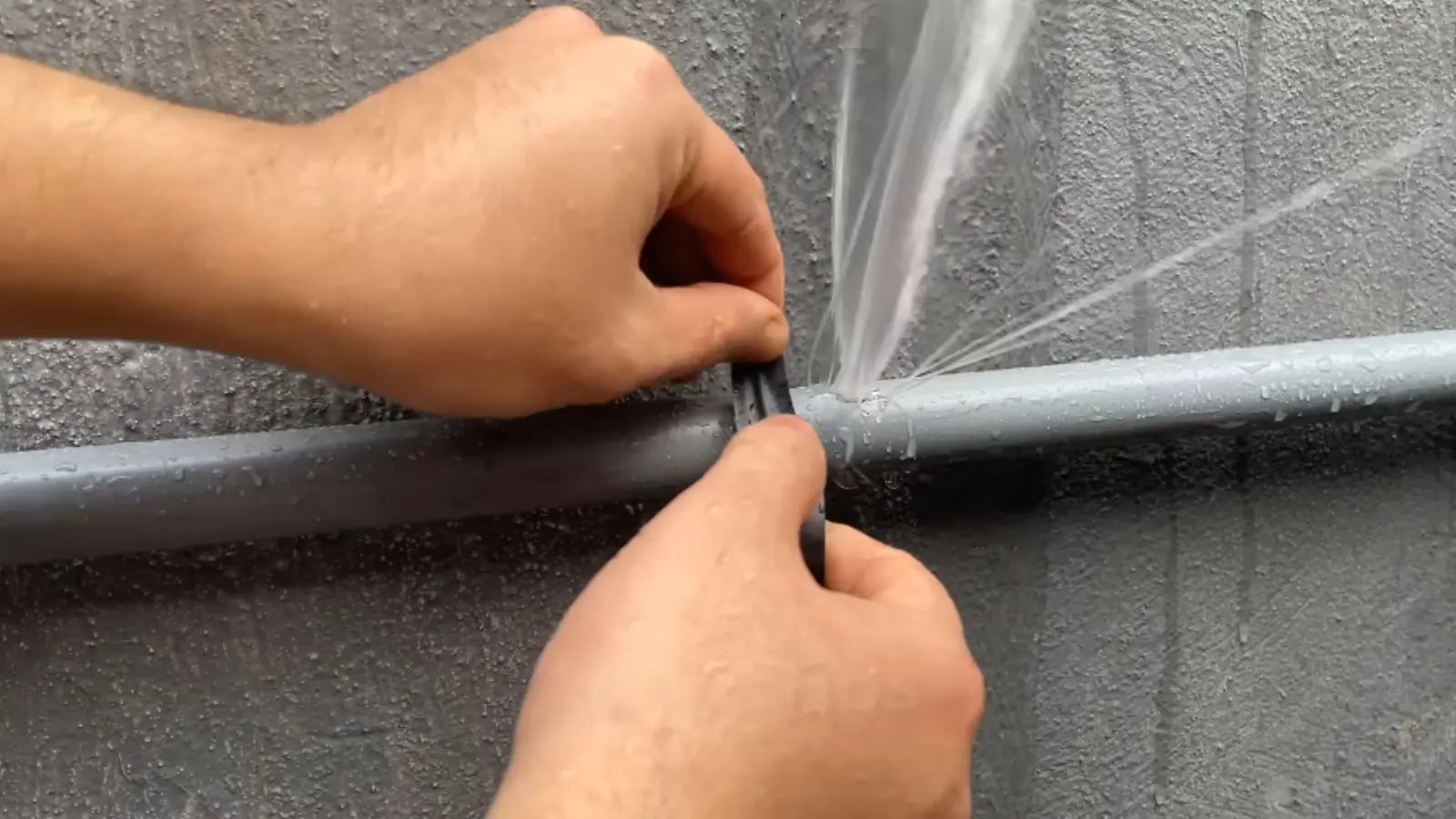 Hands clamping a leak that is spraying water from a gray pipe
