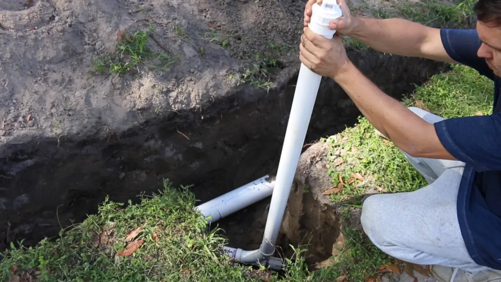Worker setting a section of white PVC sewer pipe in an open excavation